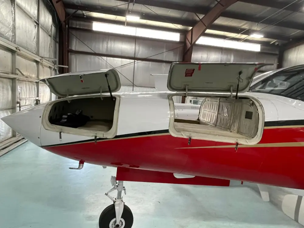 Front profile of a red and white private aircraft with its nose baggage compartments open in a hangar.