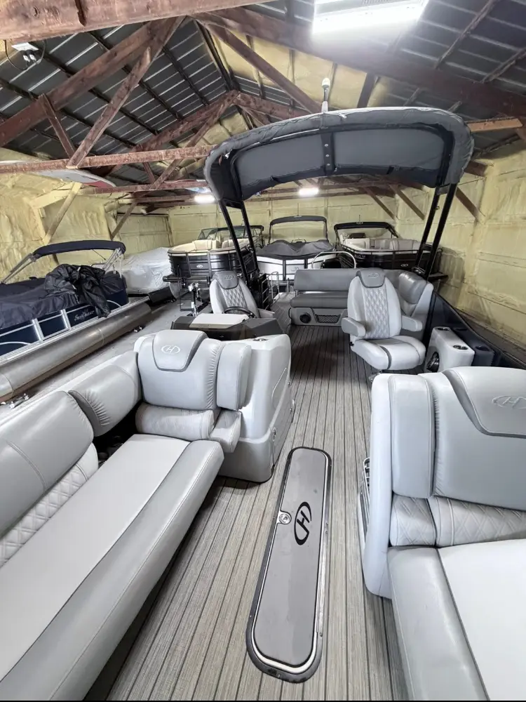 Wide interior view of a luxury pontoon boat with light gray upholstery and a bimini top, parked inside a storage shed.