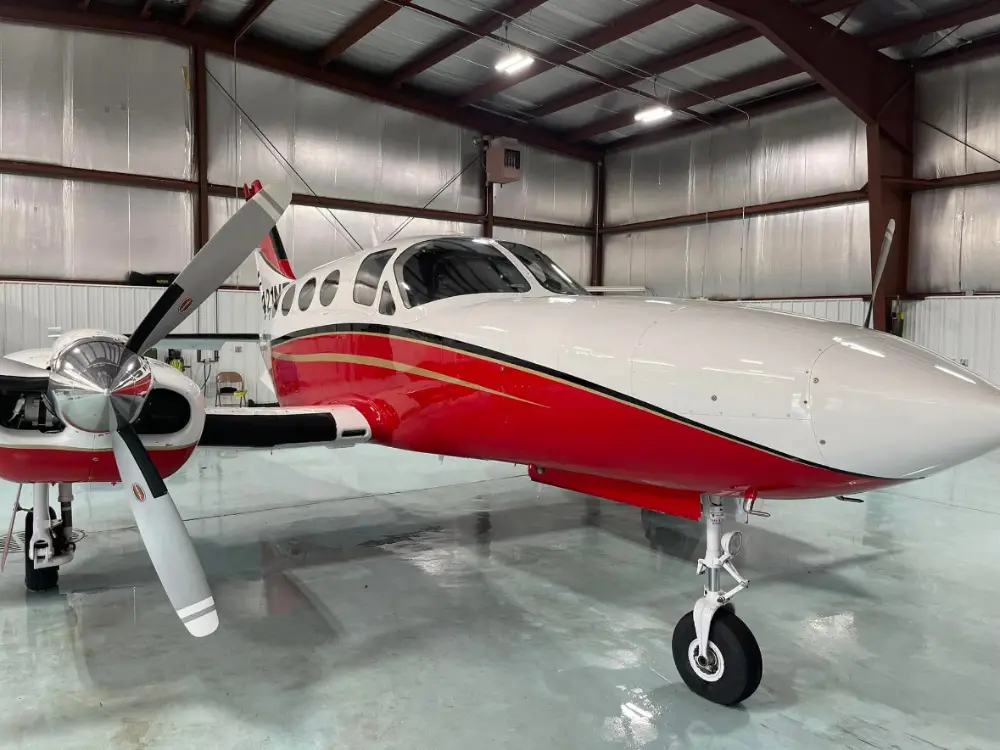 Front three-quarter view of a red and white twin-engine propeller airplane parked inside an aircraft hangar.