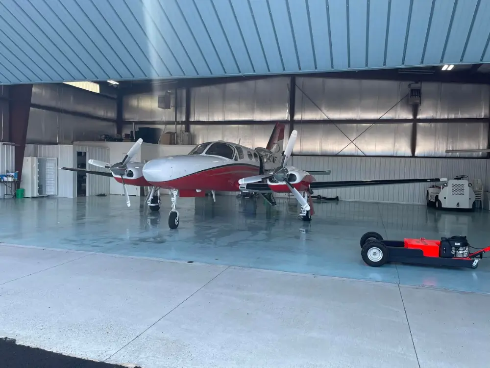 Wide shot of a red and white twin-engine airplane stored inside a large open hangar with a tug nearby.